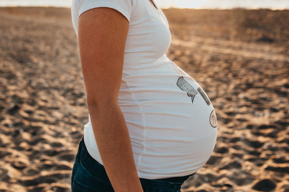 A pregnant woman standing on the beach holding her belly.