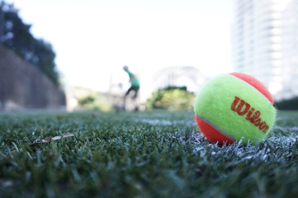 A tennis ball is in the grass with another player behind it.