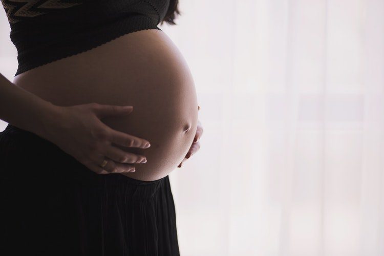 A pregnant woman is holding her belly in front of the window.